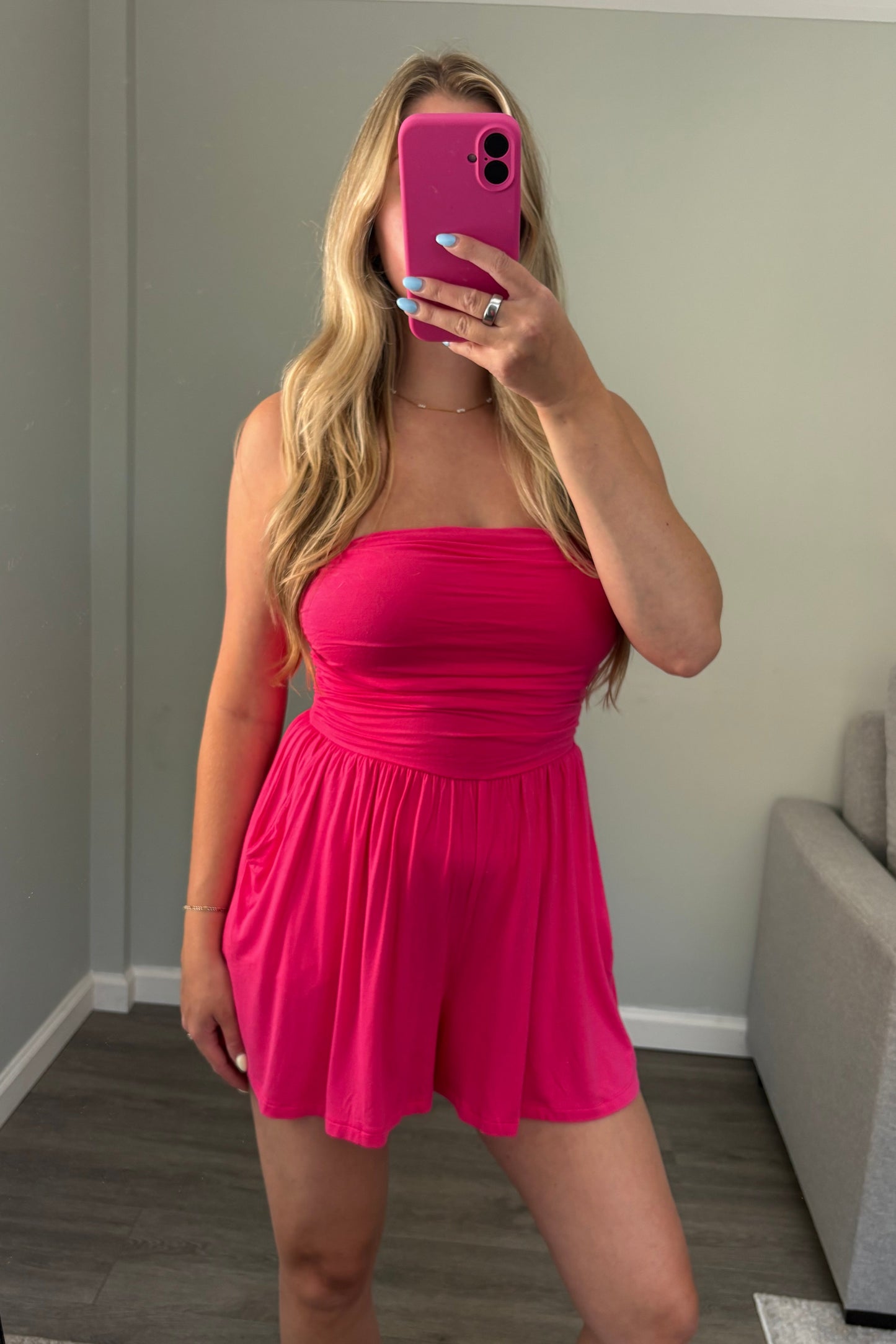 A woman takes a mirror selfie wearing a bright hot pink strapless romper in a home setting with a gray wall, wood floor, and a gray sofa in the background.