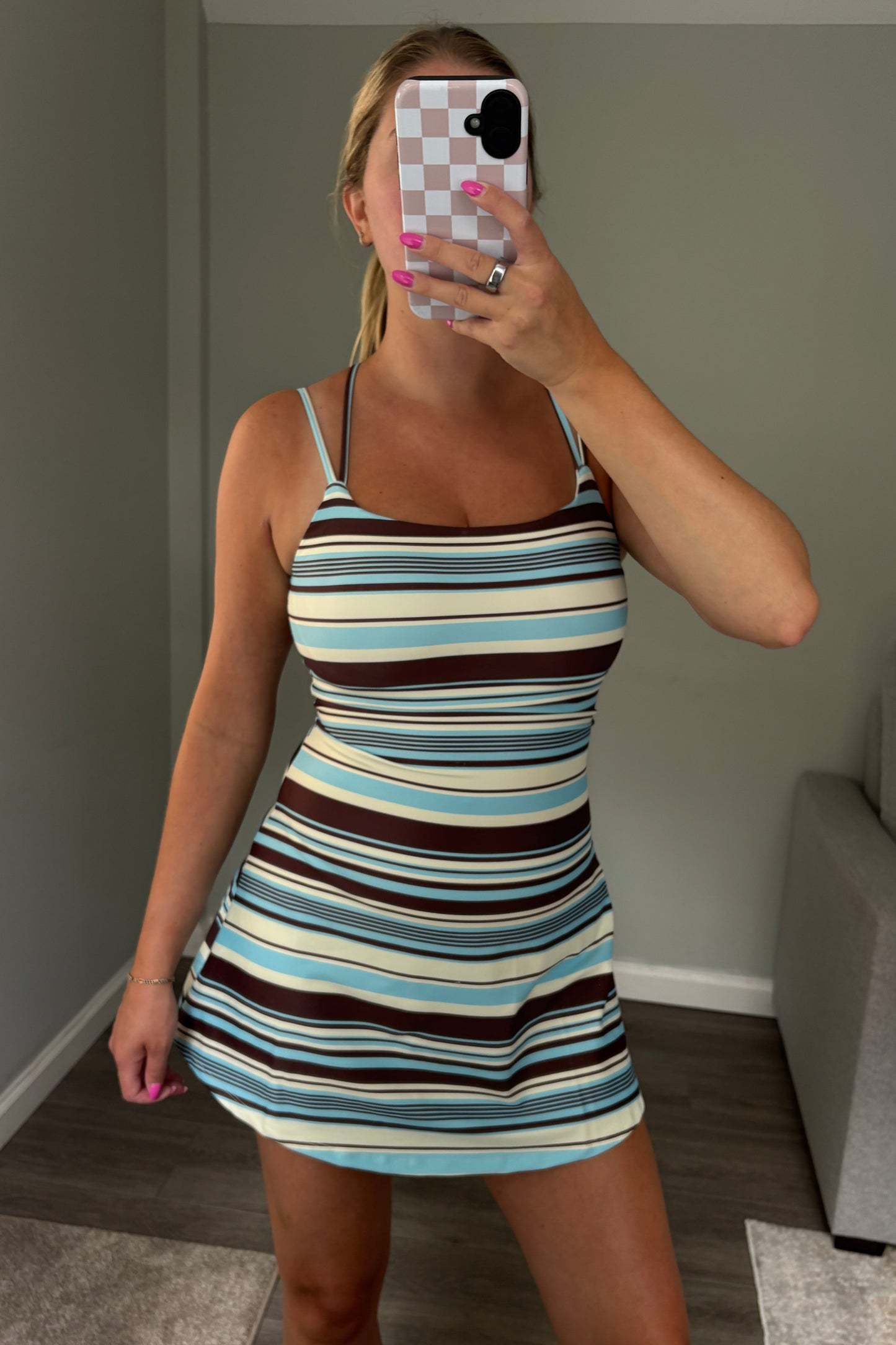 A woman takes a mirror selfie wearing a fitted mini dress with horizontal stripes in light blue, brown, cream, and black. She is standing in a room with gray walls, wood floors, and a gray sofa visible in the background.