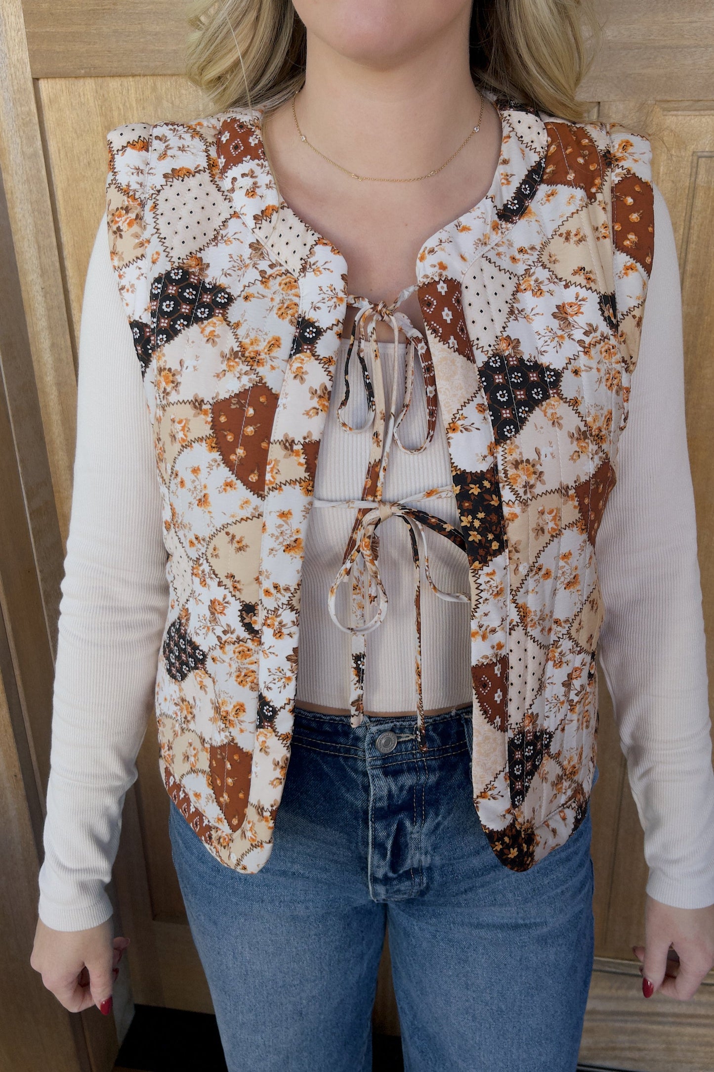 A woman wearing a quilted vest with a patchwork floral print, in shades of amber and beige, paired with a white long-sleeve top and blue jeans.