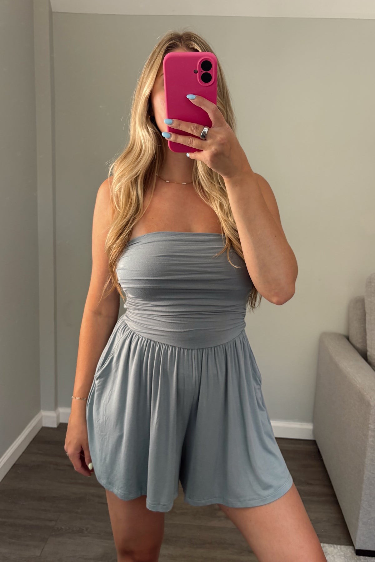 A woman takes a mirror selfie wearing a light blue-gray strapless romper in a home setting with a gray wall and a gray sofa in the background.