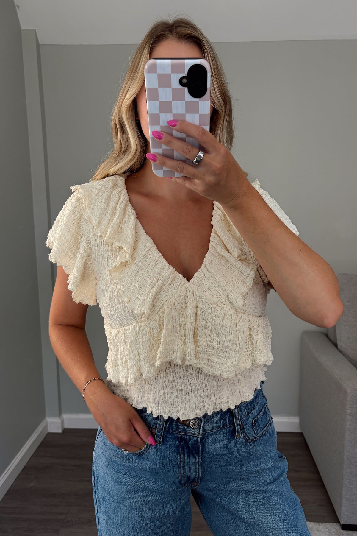 A woman takes a mirror selfie wearing a cream ruffled knit top and blue jeans in a room with gray walls and a gray sofa.