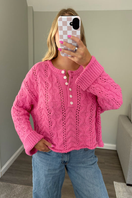 A woman takes a mirror selfie wearing a bright pink cable-knit cardigan sweater with white buttons, paired with light blue jeans, in a home setting.