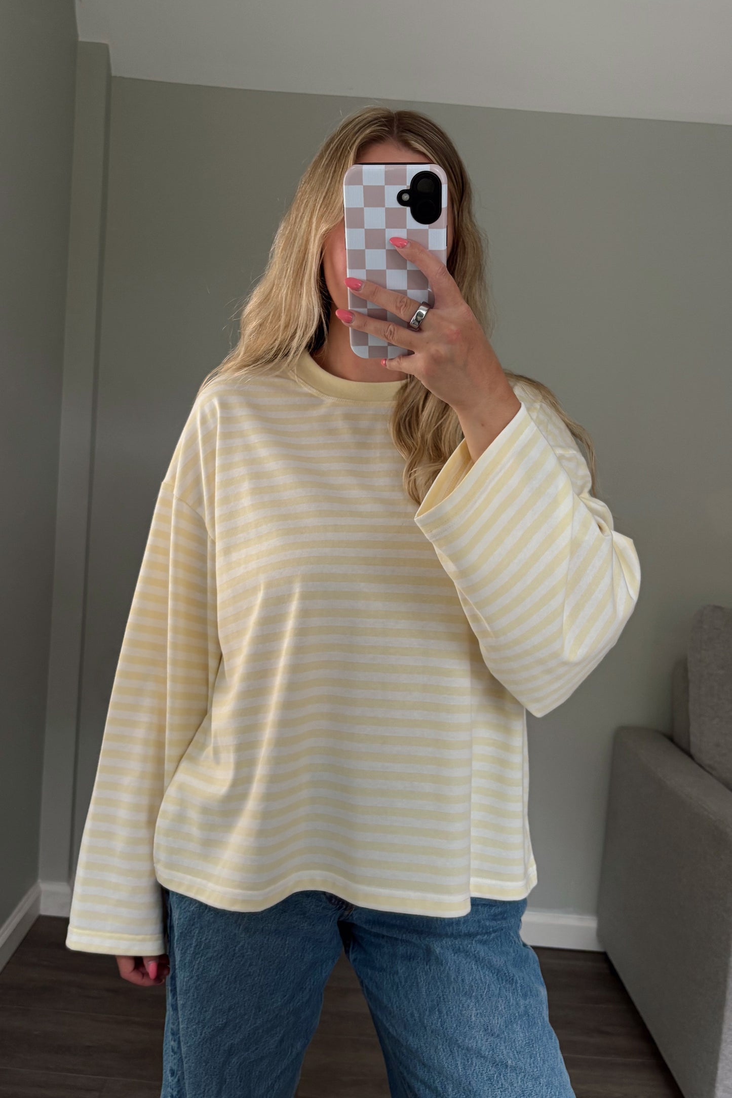 Person wearing a yellow and white striped shirt and blue jeans taking a mirror selfie in a room with a gray couch.