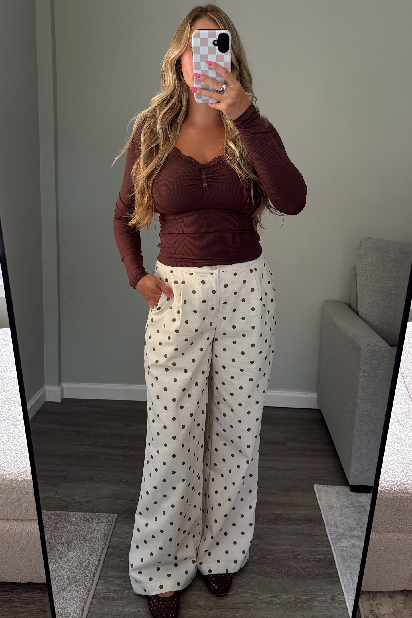 A woman takes a mirror selfie in a room, wearing a dark brown long-sleeve top and white wide-leg pants with small black polka dots. The background shows a gray wall, a gray armchair, and a large floor mirror.