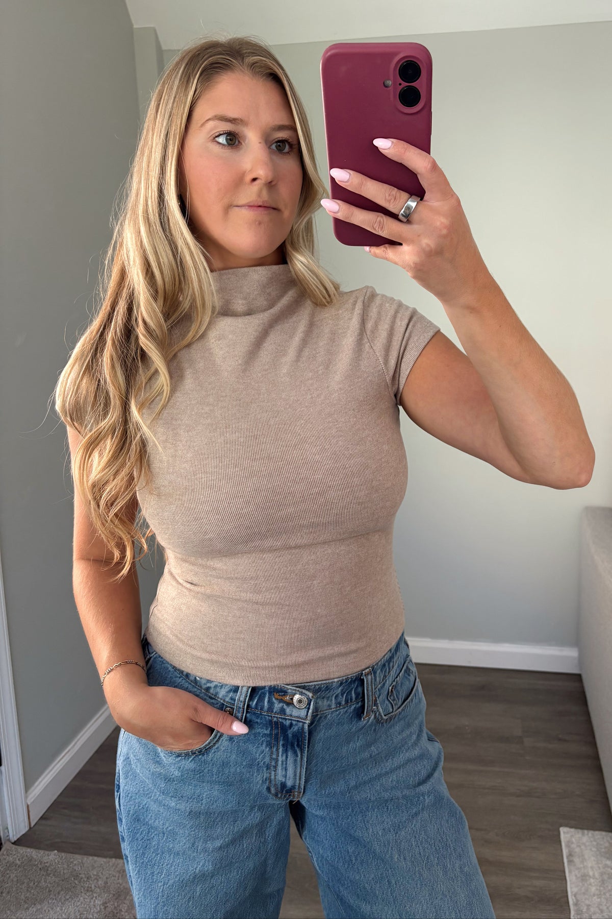 A woman taking a mirror selfie wearing a beige short-sleeve mock neck top and blue jeans in a home setting.