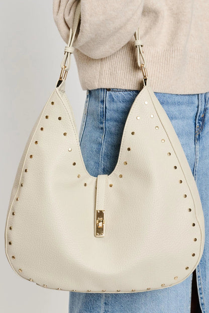 Beige handbag with gold studs held by a person wearing a beige sweater and blue jeans.