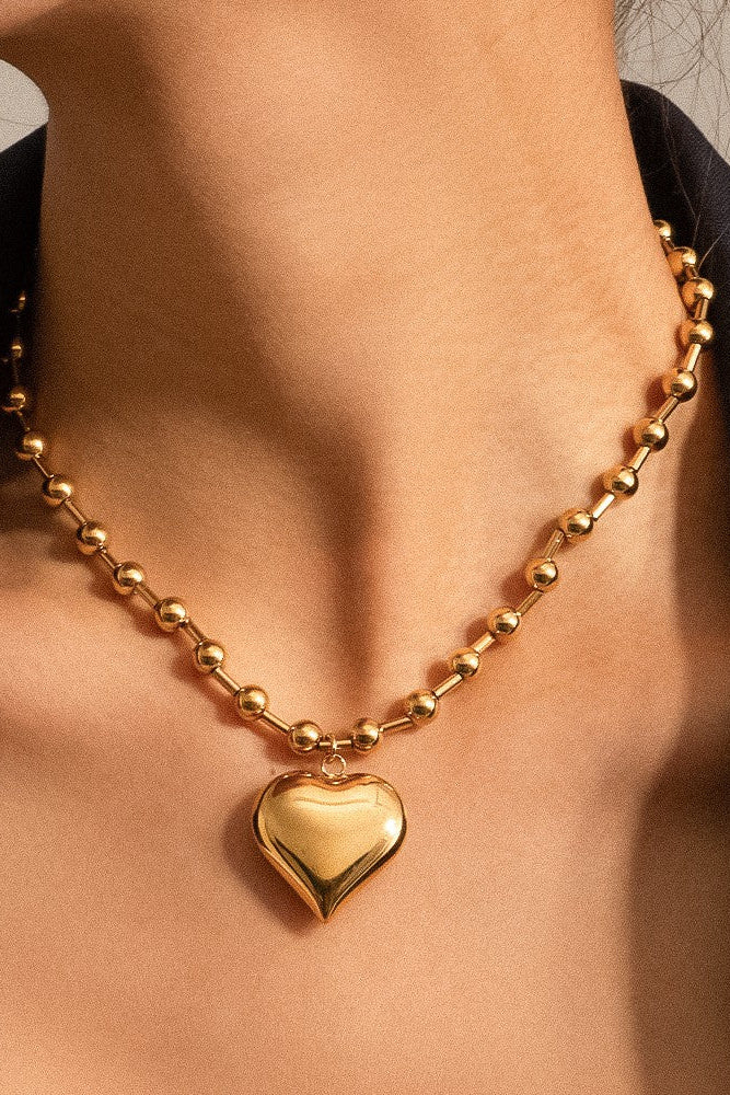 Close-up of the Paris gold-tone necklace worn around the neck, showing a polished heart pendant and beaded chain.

