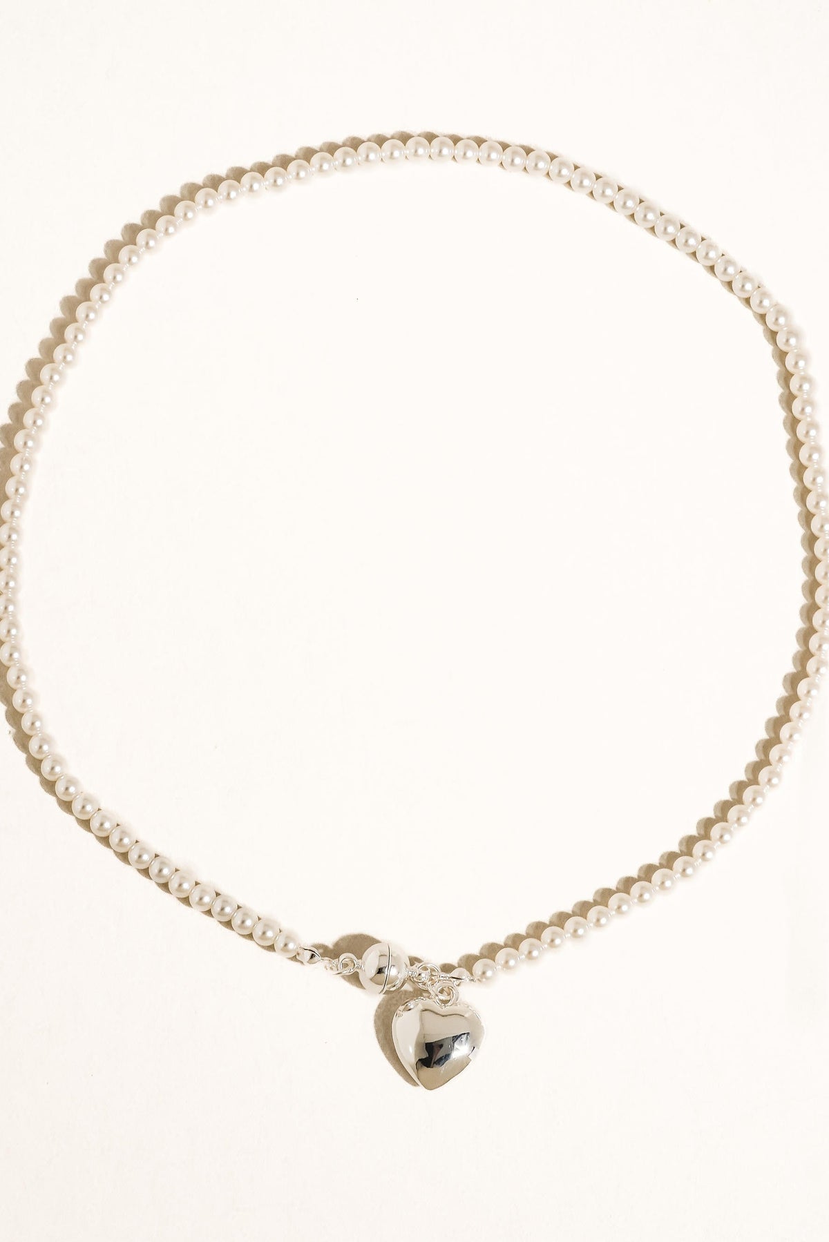 Gemma necklace arranged on a cream background, showing small pearl-style beads and a polished silver-tone heart pendant.

