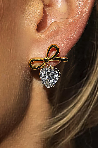 Gold bow-shaped earring with a heart-shaped gemstone worn on an ear.