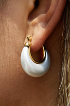 Close-up of a Roselle teardrop hoop earring worn on an ear, showing its pearly white finish and polished gold edges.

