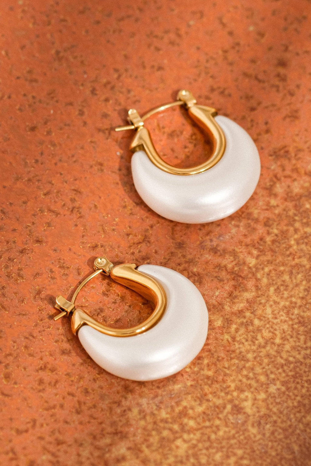 Pair of Roselle white and gold teardrop hoop earrings arranged on a terracotta-colored surface, showing their curved shape and hinged closures.

