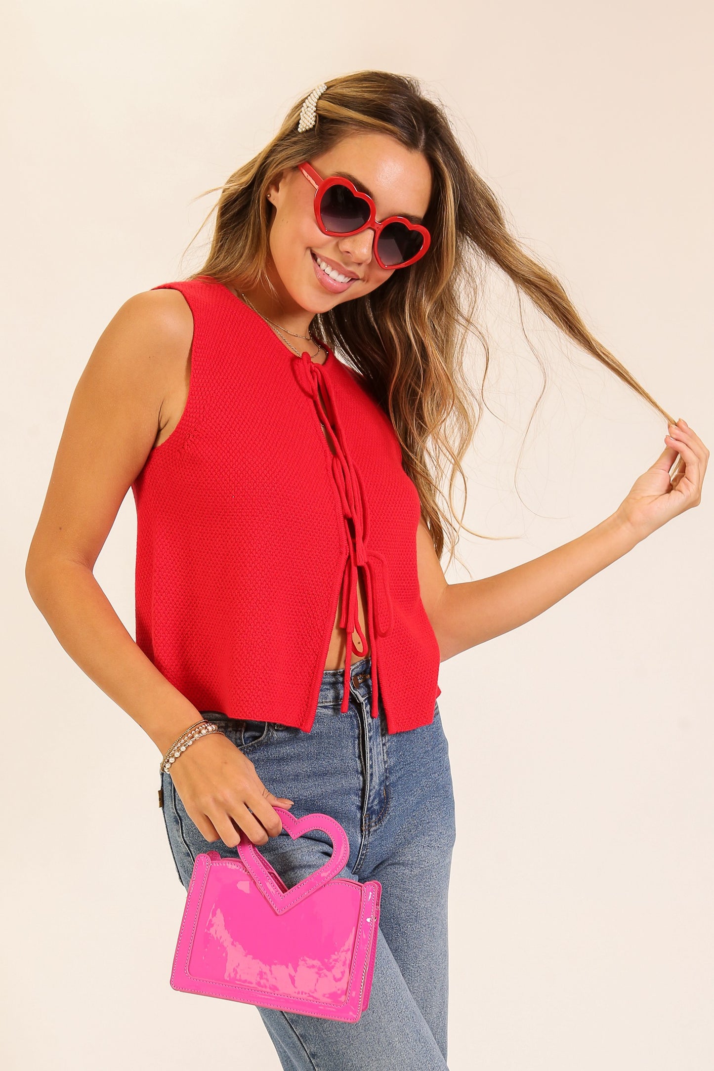Woman wearing a red sleeveless top and blue jeans, holding a pink handbag against a beige background