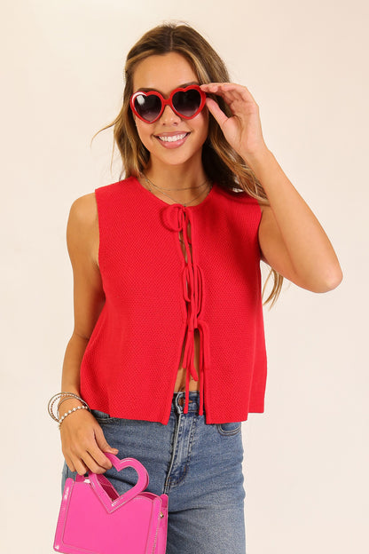 Woman wearing a red sleeveless top and blue jeans, holding pink sunglasses and a matching pink handbag.