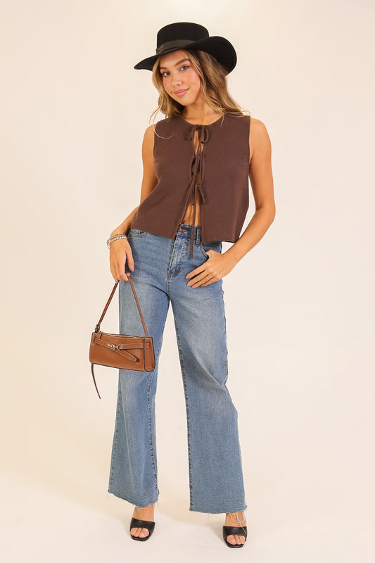 Woman wearing a brown sleeveless top, blue jeans, and a black hat, holding a brown handbag on a beige background.