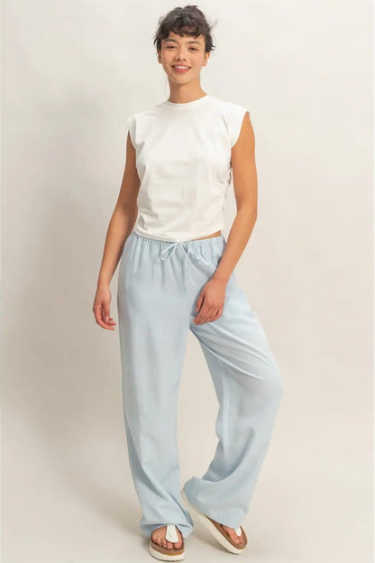 A woman modeling a sleeveless white crop top and light blue wide-leg drawstring pants against a plain beige studio background.