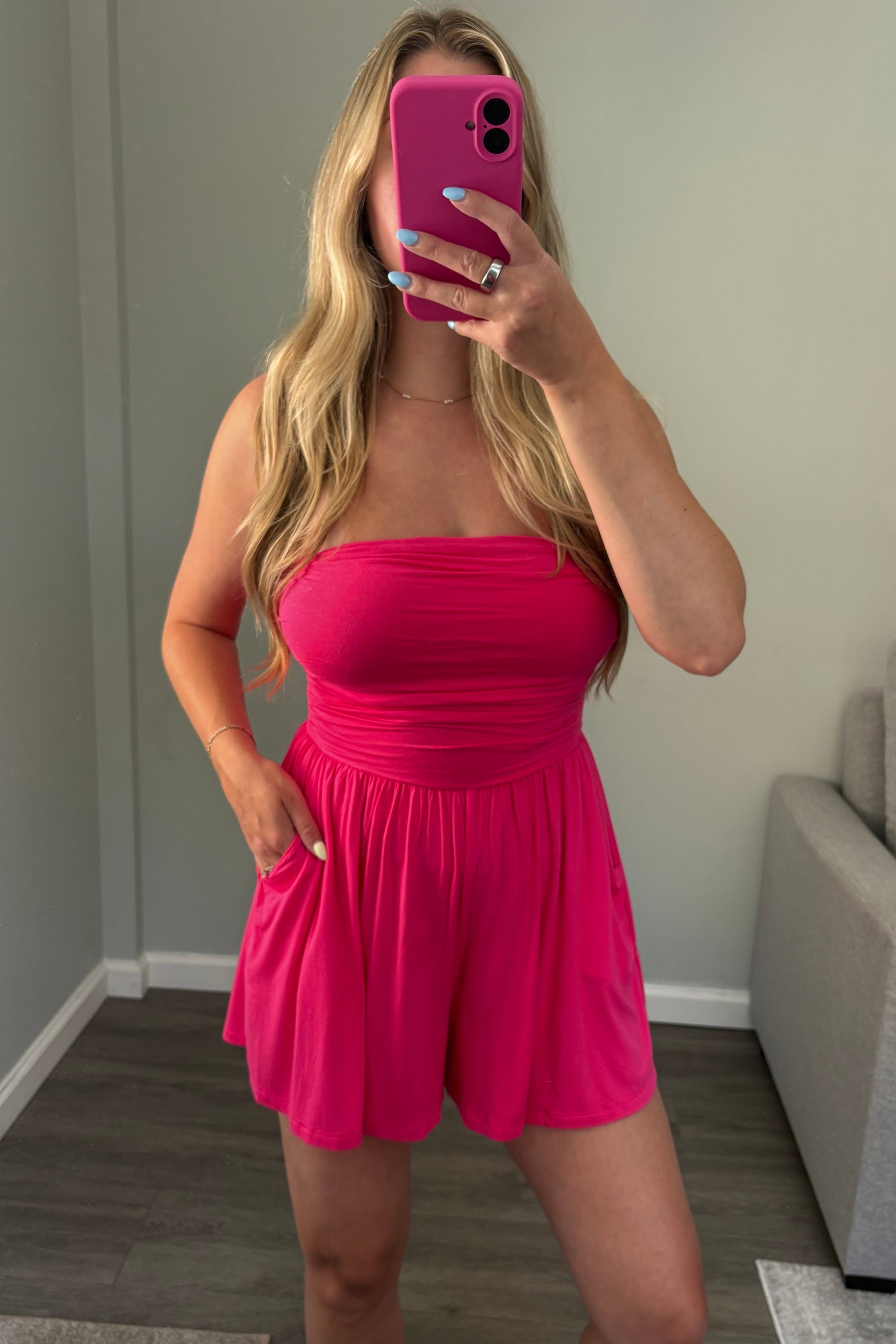 A woman takes a mirror selfie wearing a bright hot pink strapless romper with side pockets, standing in a room with gray walls and wood floors.