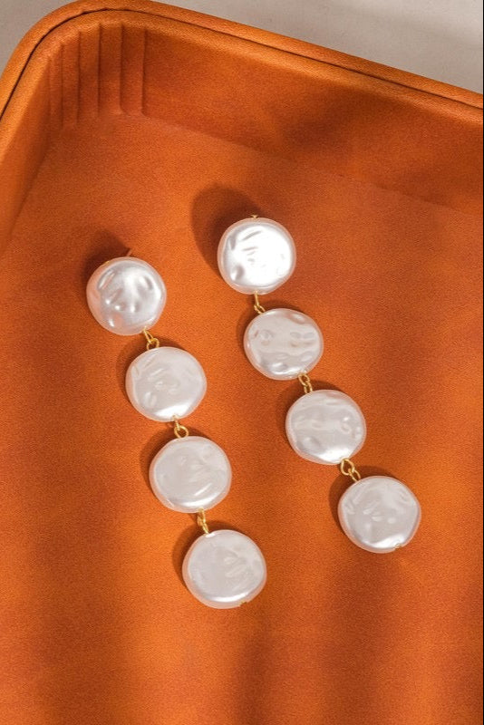 Heloise faux pearl drop earrings laid flat on an orange-brown tray, each with four lustrous white discs and gold-tone connecting links.

