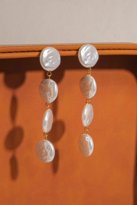 Pair of Heloise faux pearl drop earrings hanging from the edge of an orange-brown tray, showing their tiered design.

