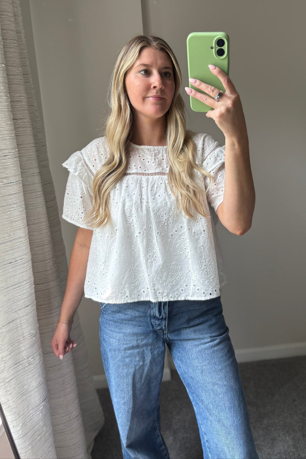 Front mirror view of the Blossom white eyelet blouse with blue jeans, showing its loose shape and straight hem.

