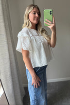 Side mirror view of the Blossom white eyelet blouse showing its layered sleeve ruffle and relaxed fit over blue jeans.

