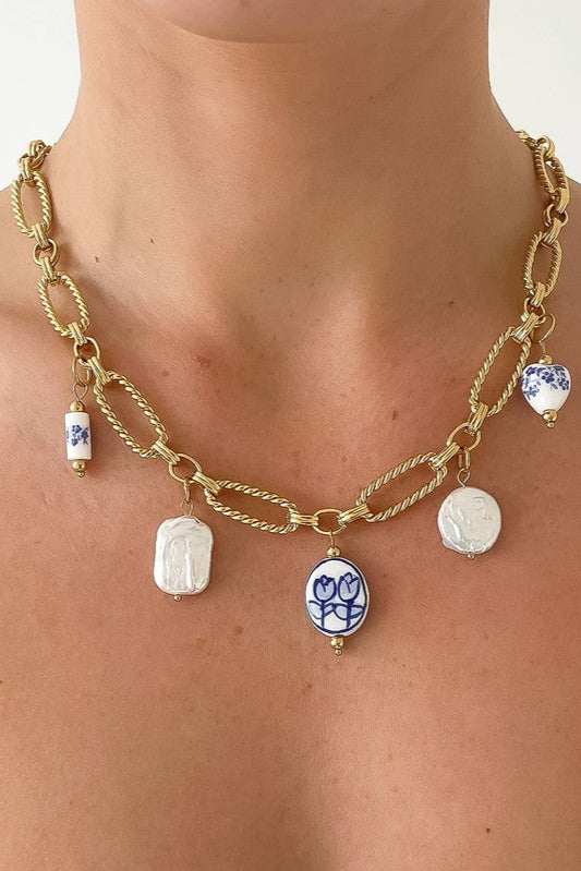 Riela gold-tone chain necklace worn around the neck, with blue-and-white floral ceramic charms and white faux pearl accents.

