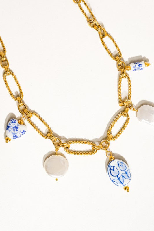 Close-up of the Riela necklace showing blue floral ceramic charms, white faux pearls, and textured gold-tone chain links.

