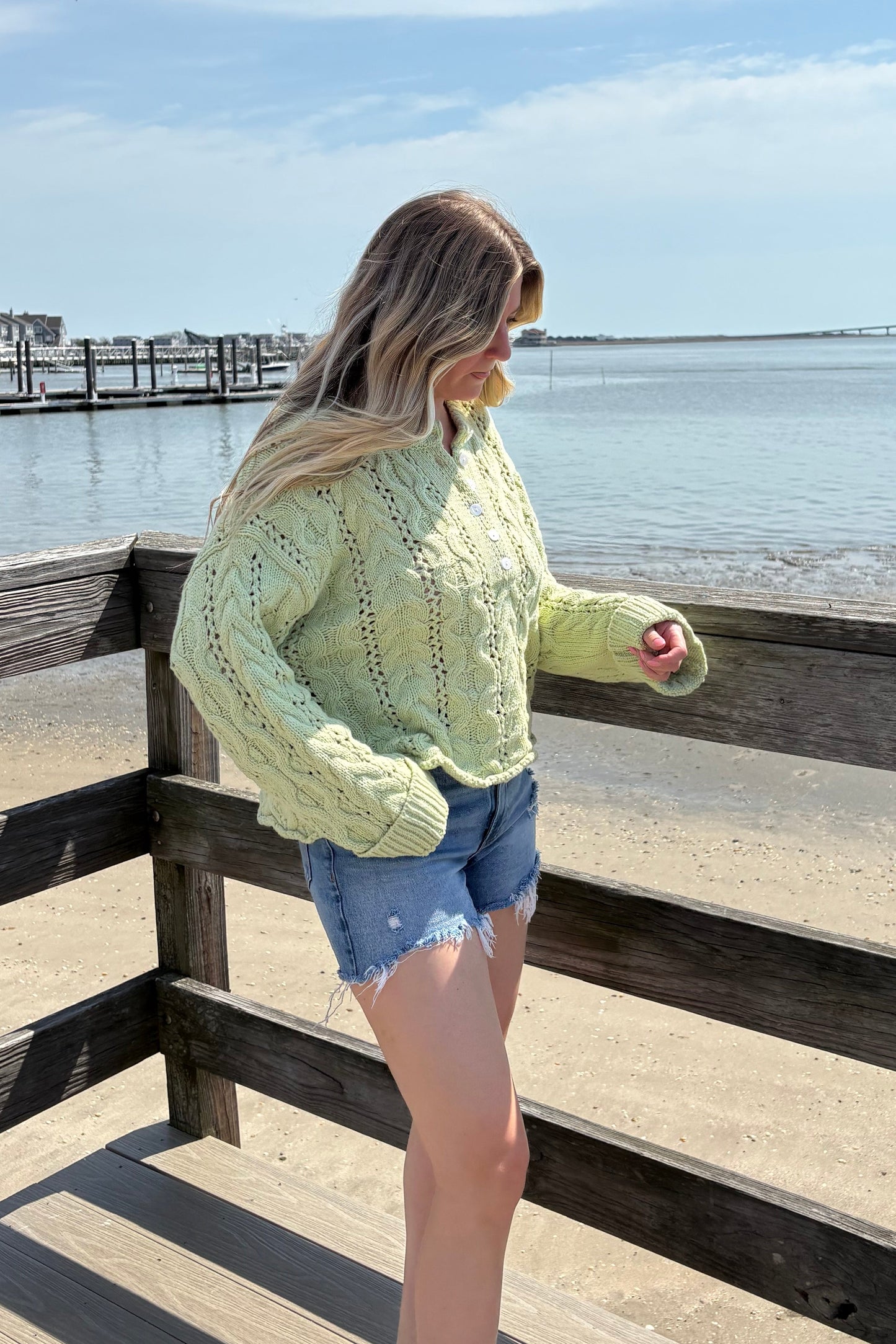 Person wearing a light green cardigan on a wooden deck with a clear blue sky.