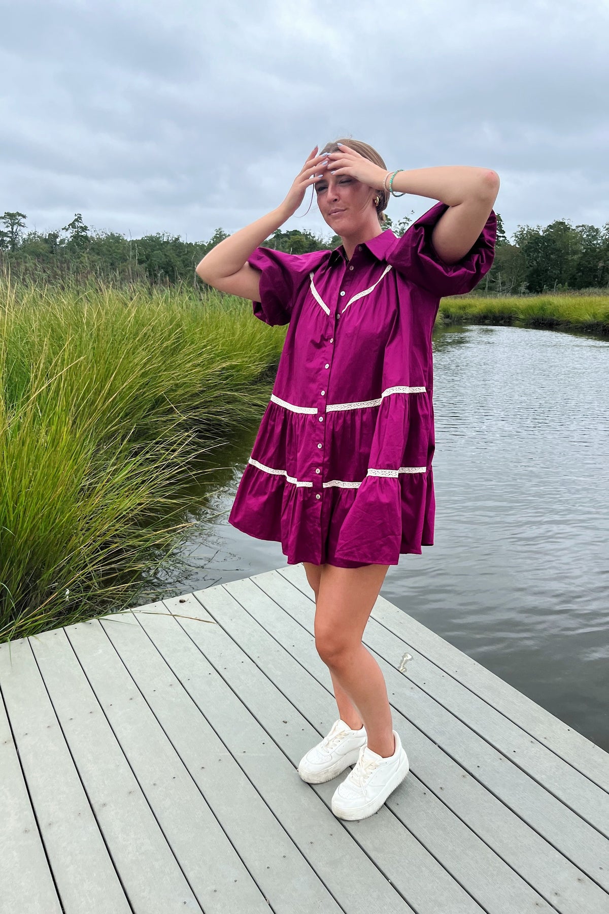 Model on a dock wearing a magenta button-front mini dress with puff sleeves, white crochet trim, and white sneakers.

