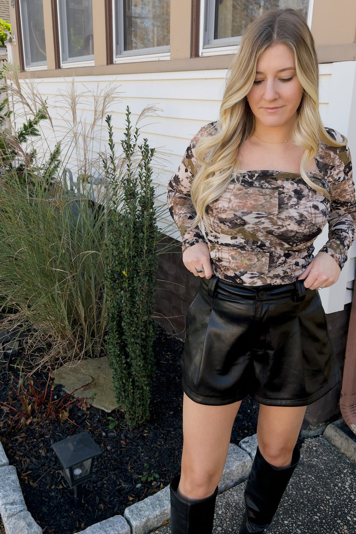Model holding the waistband of Nina black faux leather shorts, showing the front pleats and curved hems.

