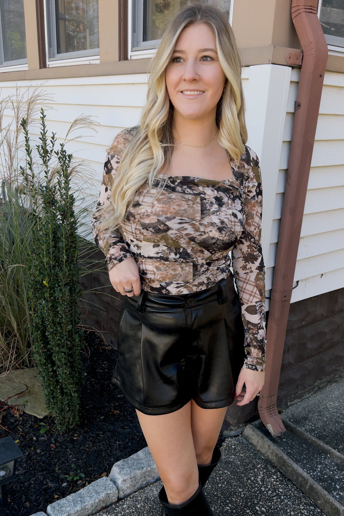 Model wearing Nina black faux leather shorts with front pleats, styled with a floral long-sleeve top and black boots.

