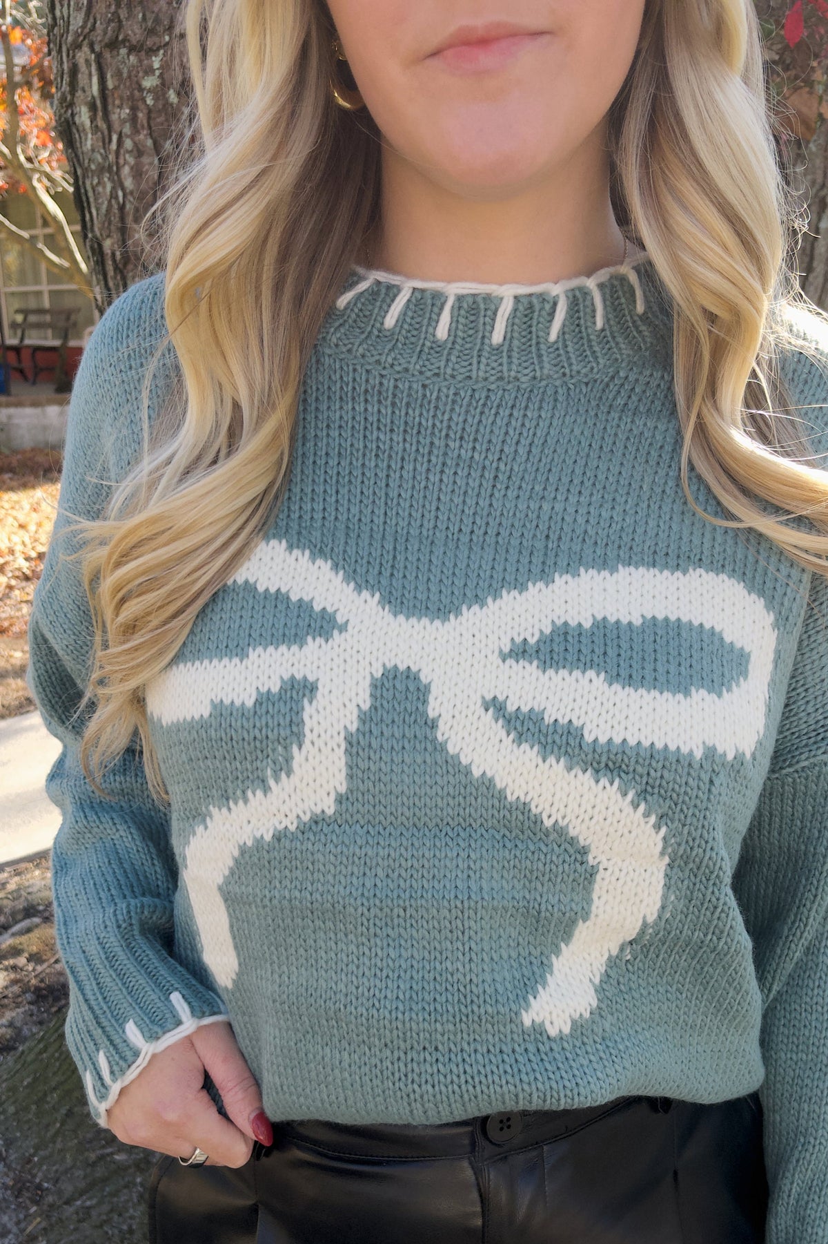 Close-up of the Vivian sweater showing the cream bow graphic, blue knit texture, and contrast stitching at the crew neckline.

