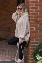 Side view of a model wearing the sand beige oversized knit sweater with black leggings and holding a beige shoulder bag.


