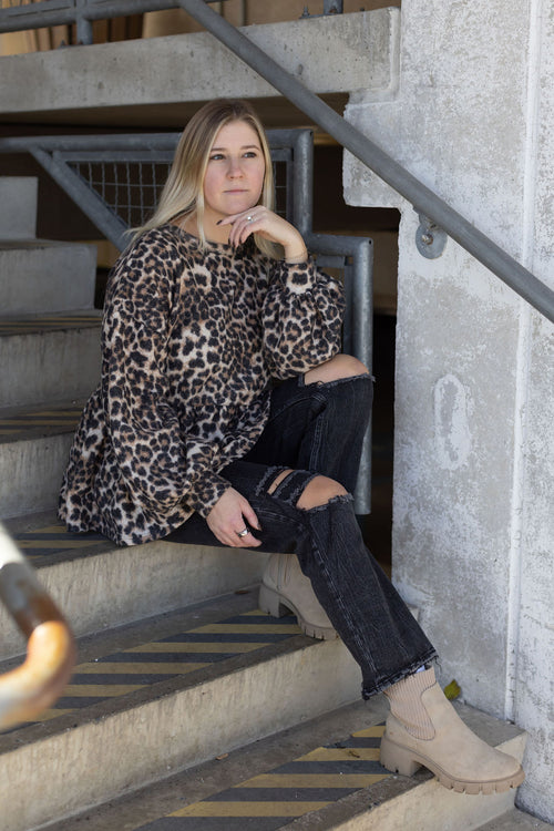 Model seated on concrete stairs wearing a leopard print babydoll top, ripped black jeans, and beige ankle boots.

