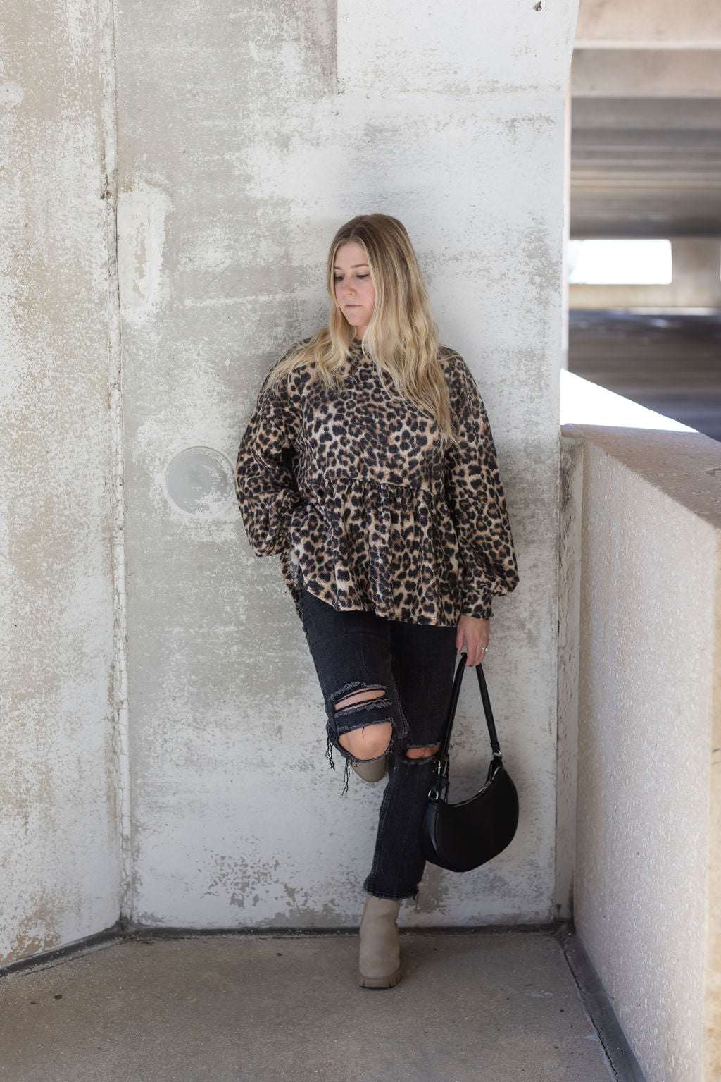 Model wearing a leopard print babydoll top with distressed black jeans, beige boots, and a black handbag.

