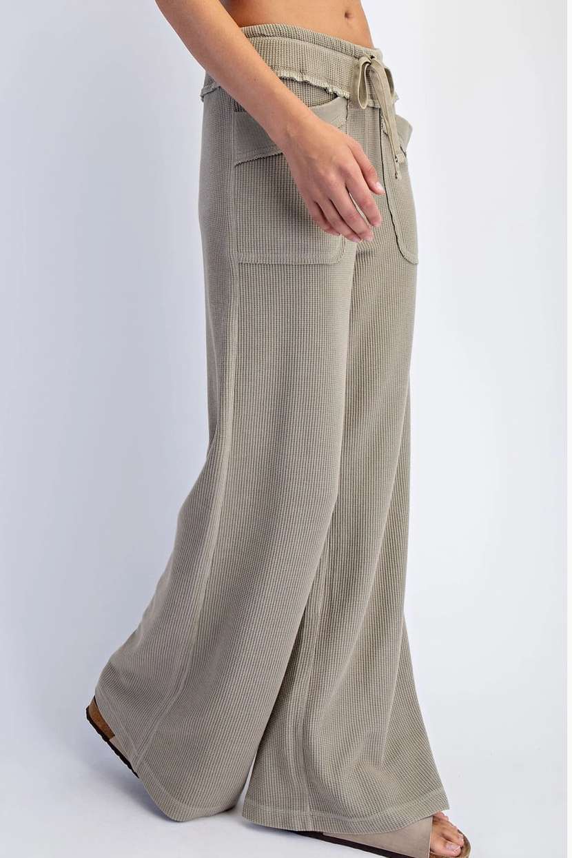 A person wearing wide-leg pleated knit pants in a light taupe-beige color, paired with brown flat sandals, shown from the waist down against a light gray studio background.
