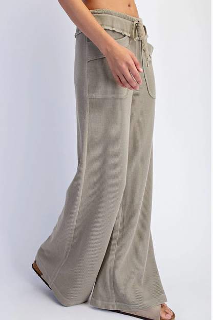 A person wearing wide-leg pleated knit pants in a light taupe-beige color, paired with brown flat sandals, shown from the waist down against a light gray studio background.