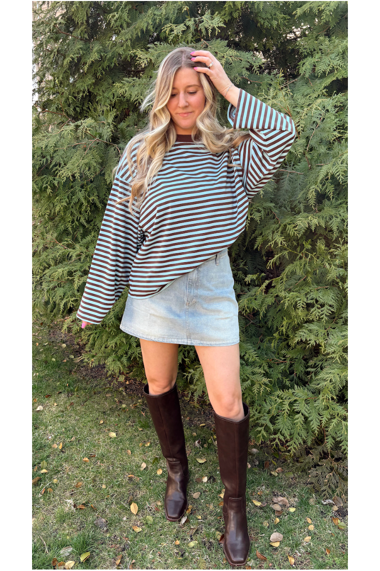 Woman wearing a striped top, denim skirt, and knee-high boots standing in front of green trees.