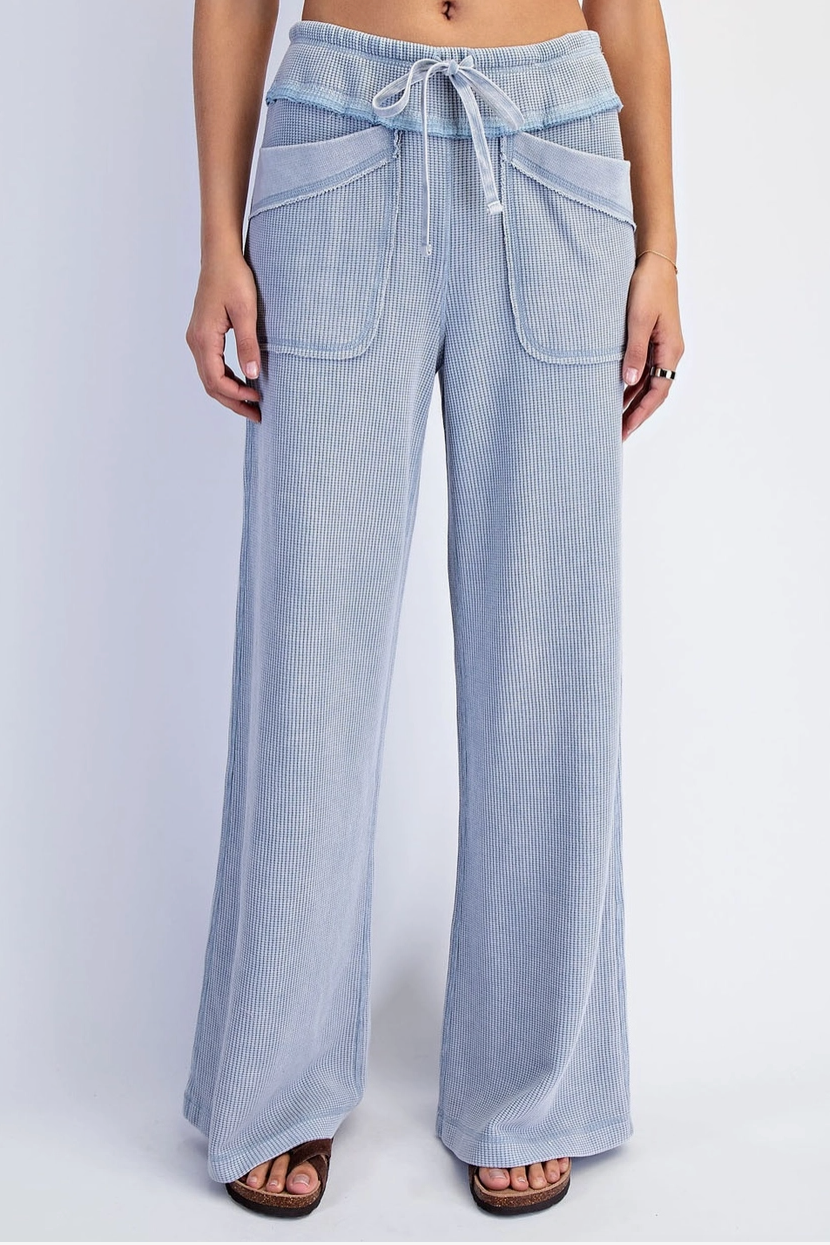 A model is wearing light blue wide-leg pants with a drawstring waist and large front patch pockets, paired with brown flat sandals against a light gray studio background.