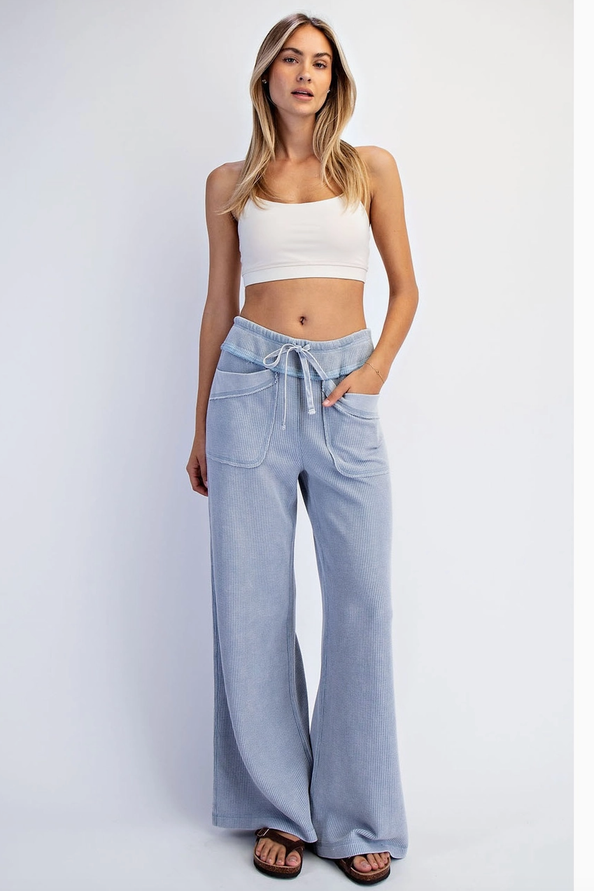 A woman modeling a white ribbed crop top and light blue wide-leg drawstring pants against a plain light gray studio background.