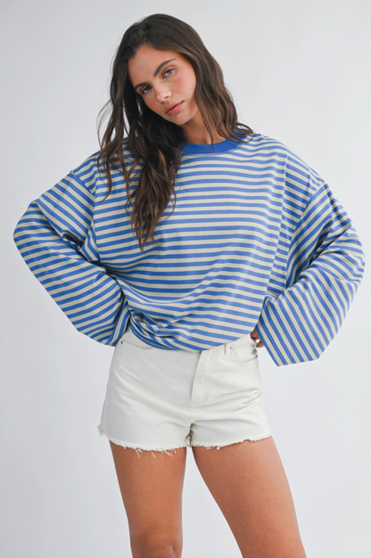 A woman modeling a blue and white striped long-sleeve t-shirt with balloon sleeves, paired with white denim shorts, against a light gray studio background.