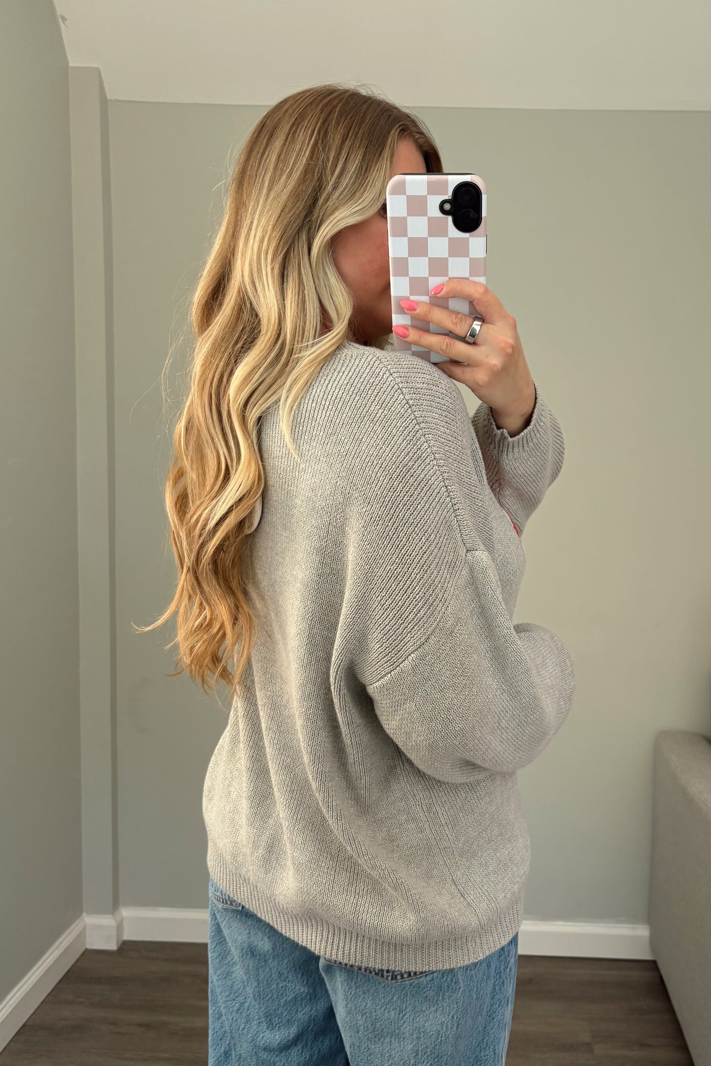 Woman taking a mirror selfie wearing a gray sweater and blue jeans in a room with a couch and window.