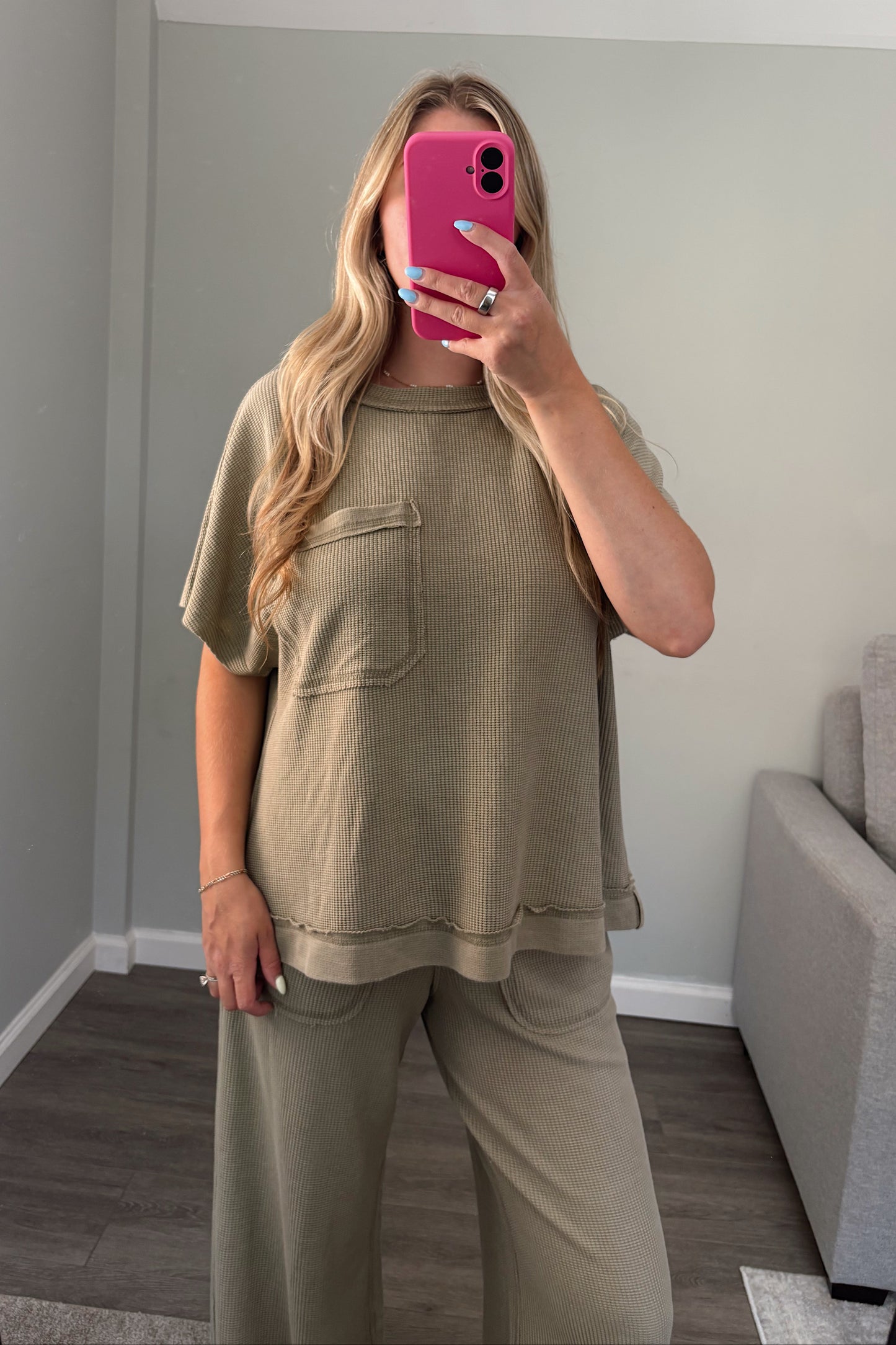 A woman takes a mirror selfie wearing a matching two-piece lounge set in a muted olive green color, consisting of a short-sleeve top with a chest pocket and wide-leg pants, in a modern room with gray walls and wood flooring.