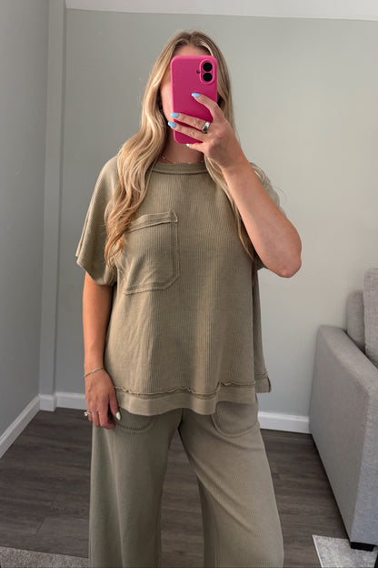 A woman takes a mirror selfie wearing a matching two-piece lounge set in a muted olive green color, consisting of a short-sleeve top with a chest pocket and wide-leg pants, in a modern room with gray walls and wood flooring.