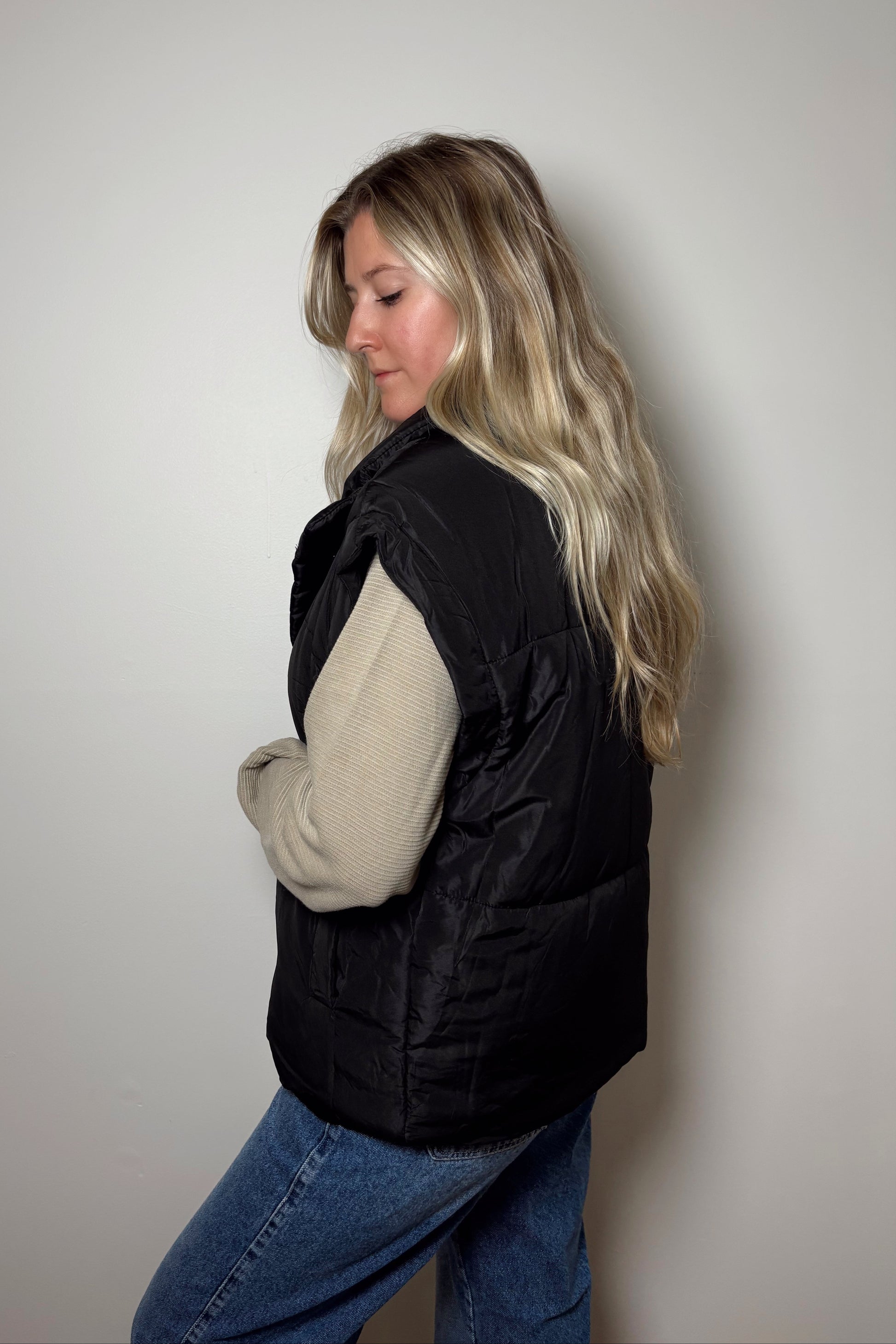Woman wearing a black puffer vest over a beige sweater with blue jeans against a plain background