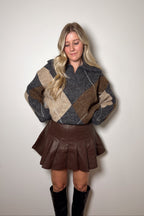 Harper brown faux leather pleated mini skirt styled with an argyle sweater and tall black boots.

