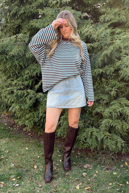 Person wearing a striped sweater and denim skirt standing in front of evergreen trees.