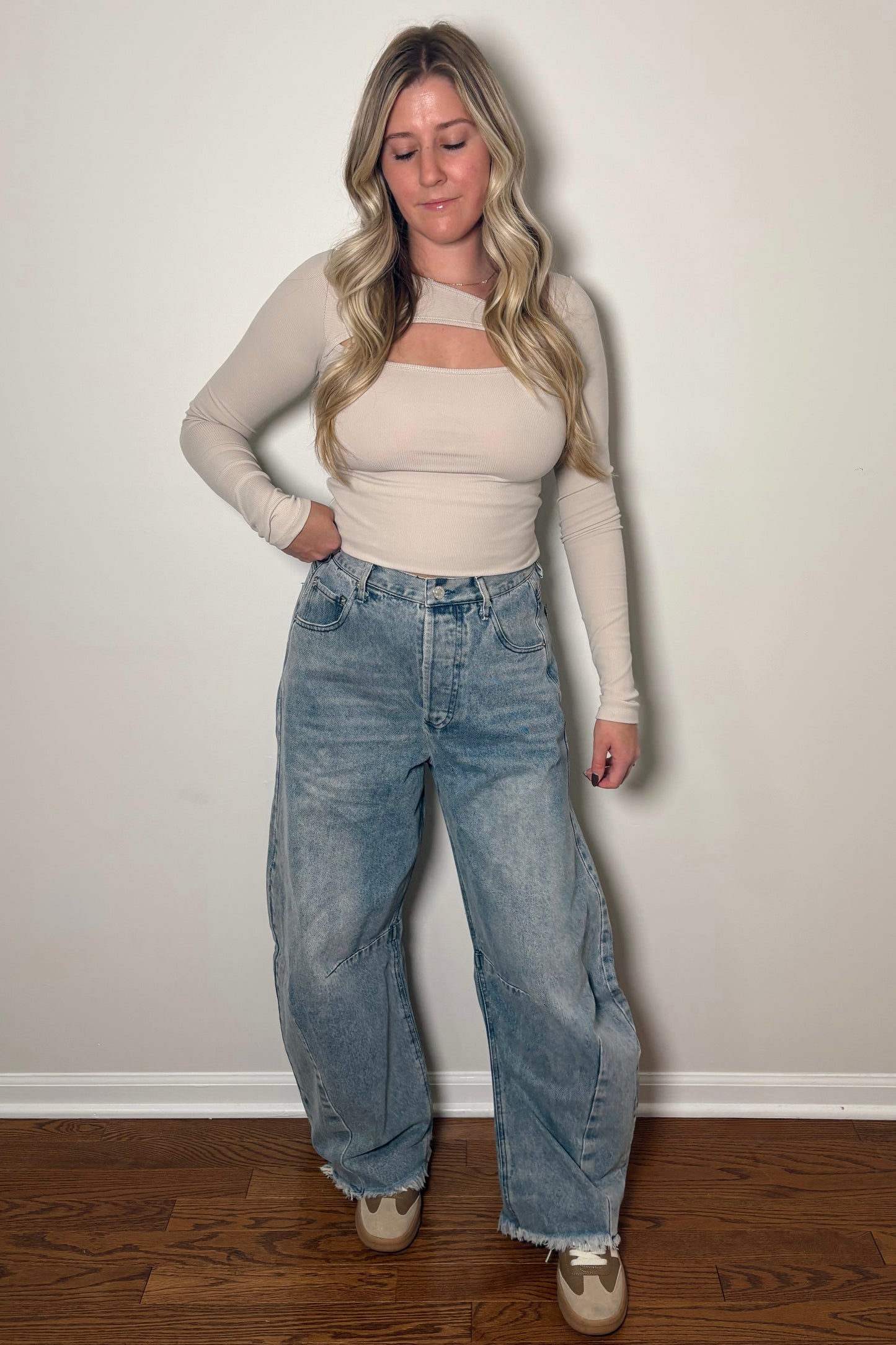 Woman wearing a beige long-sleeve top and blue jeans against a plain white wall.