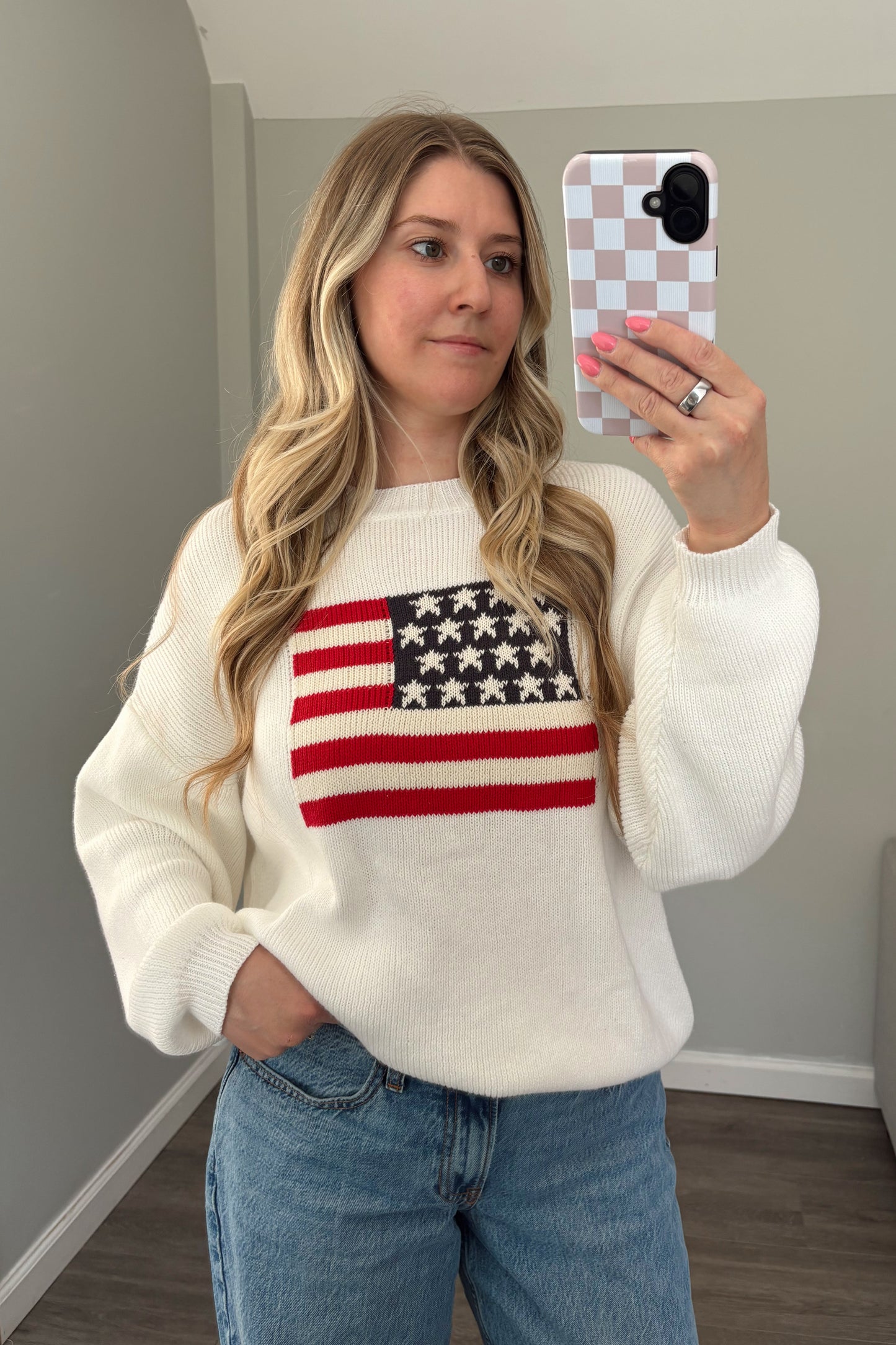 Woman wearing a white sweater with an American flag design, taking a mirror selfie.