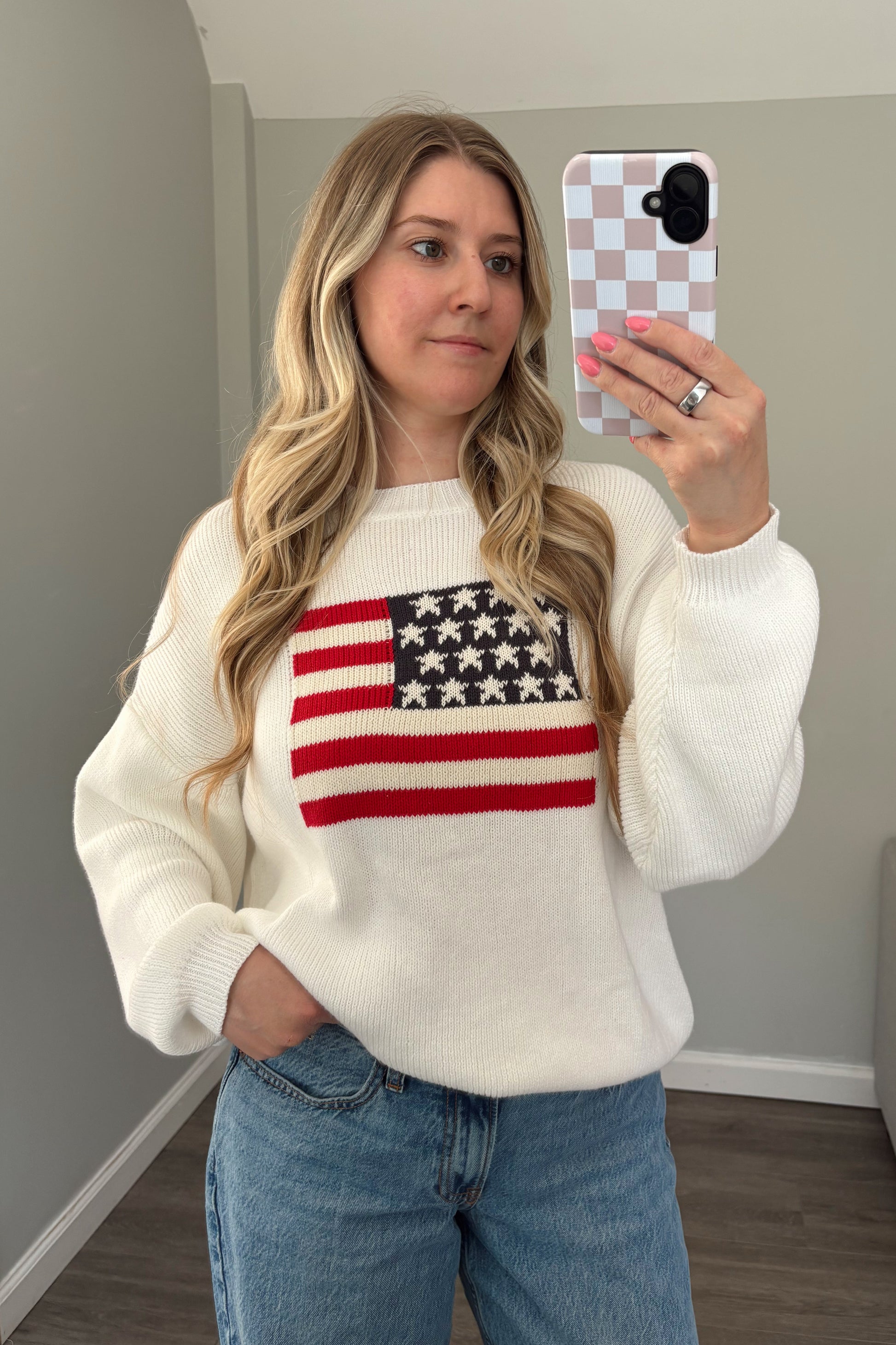 Woman wearing a white sweater with an American flag design, taking a mirror selfie.
