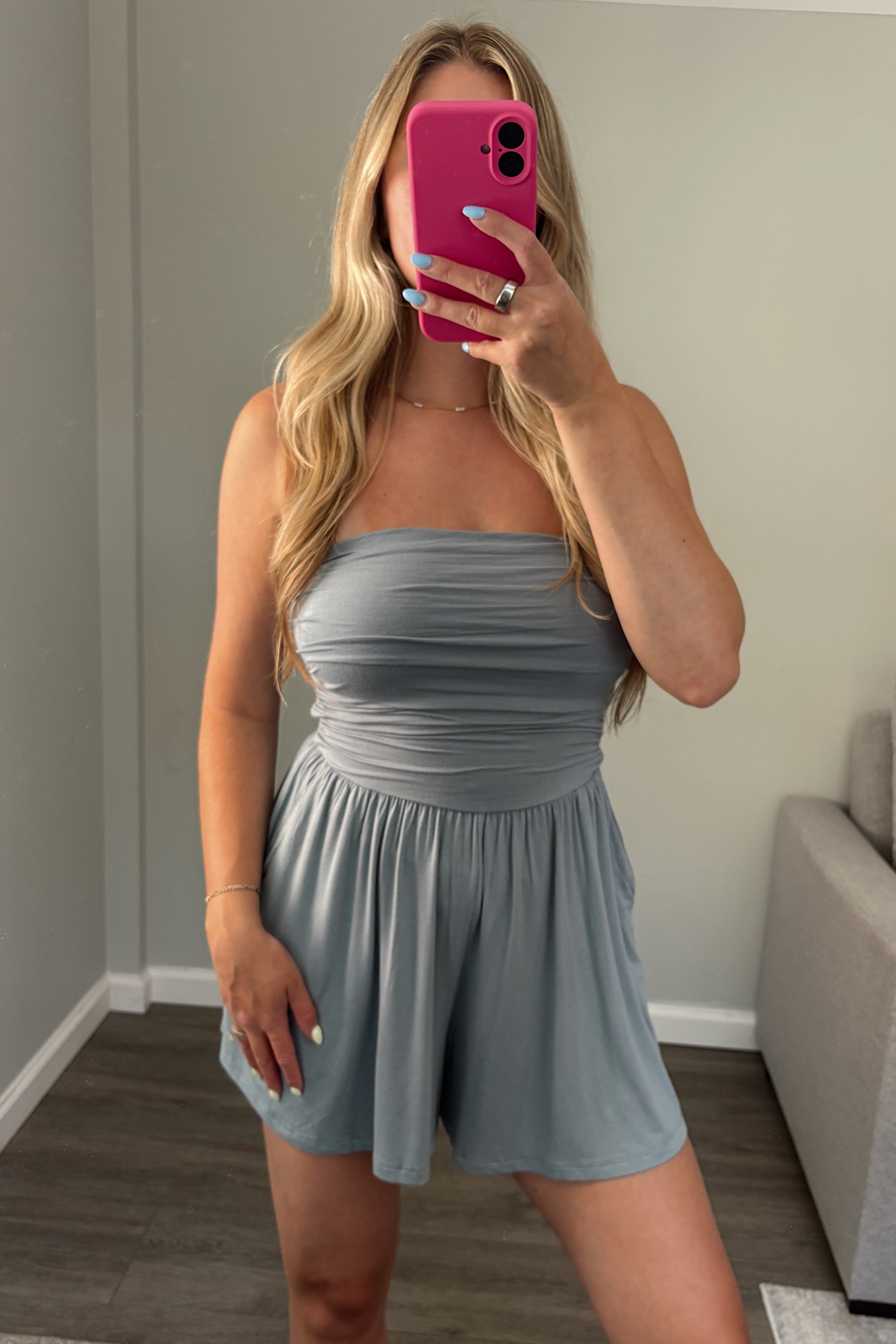 A woman takes a mirror selfie wearing a light blue-gray strapless romper in a room with gray walls and wood-look flooring.